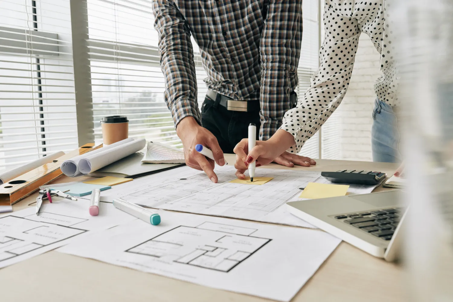 Dos personas colaborando en un proyecto de diseño arquitectónico, revisando planos en una mesa de trabajo con materiales de oficina y una computadora portátil.