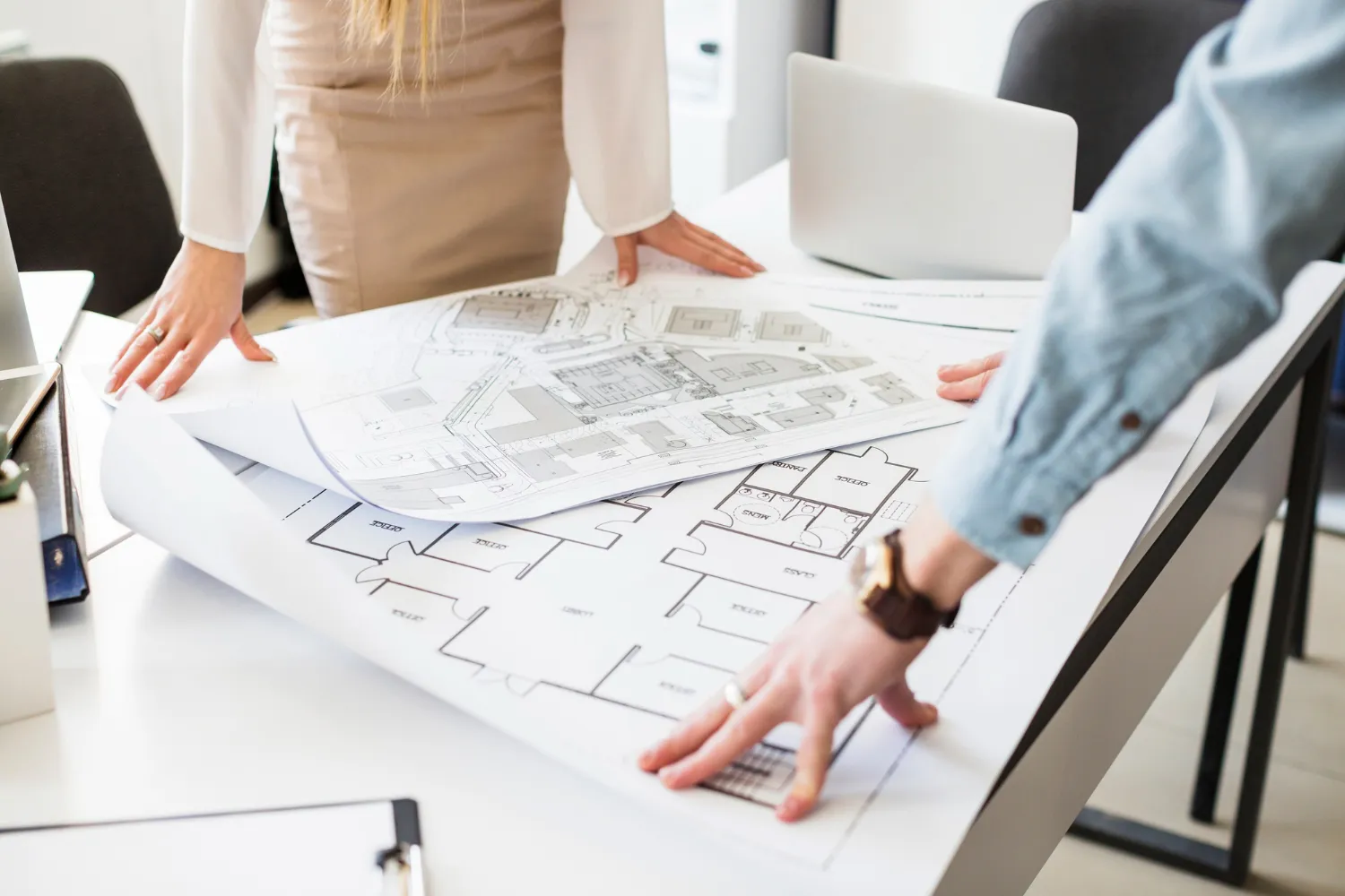 Manos de profesionales revisando planos arquitectónicos sobre una mesa, con una computadora portátil en el fondo.