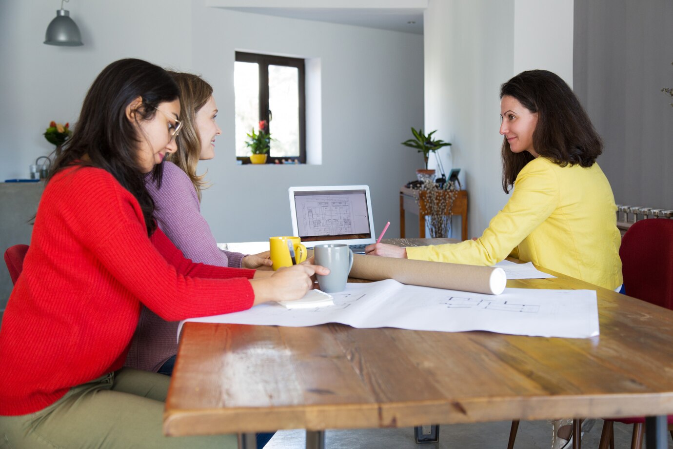 Tres mujeres colaborando en una reunión de trabajo en una mesa de madera, con un ordenador portátil y documentos sobre el diseño en un ambiente luminoso y moderno.
